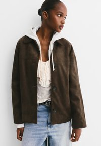 Veste en daim marron avec un col, fermeture à bouton, et un sweat à capuche gris qui dépasse en dessous. Portée sur un jean clair et un haut à volants.