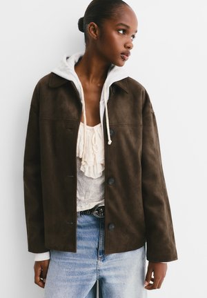 Bruin suède jack met kraag, knoopsluiting en een grijze hoodie die eronderuit piept. Draagt over lichte jeans en een gerimpeld topje.