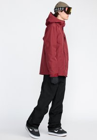 Veste imperméable rouge avec capuche, pantalon noir et baskets noires. Comprend des lunettes réfléchissantes et un bonnet texturé.