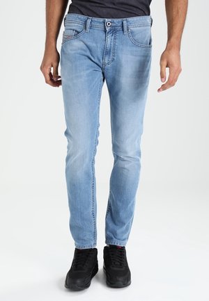 Homme portant un jean slim bleu clair et des baskets noires, debout devant un fond blanc uni.