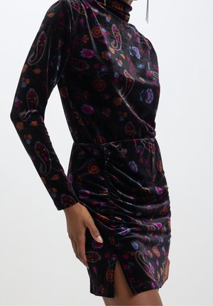 Woman wearing a long-sleeve, black velvet dress with colorful floral and paisley patterns and a ruched detail near a front slit.