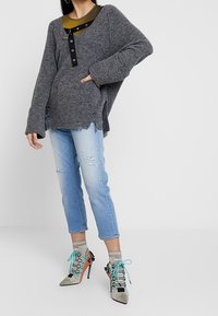 Grauer übergroßer Pullover mit geripptem Saum und schwarzem Knopfakzent. Hellblaue, distressed Jeans und silberne High Heels mit türkisfarbenen Schnürsenkeln.
