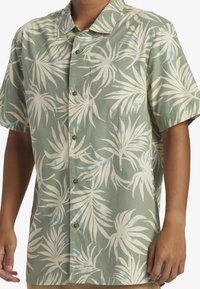 Camisa de manga corta con botones de tela verde, que presenta un patrón tropical de hojas en color crema con toques azules y botones marrones.
