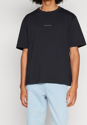 Persona che indossa una T-shirt nera a maniche corte con la scritta "ABERCROMBIE" sul petto e jeans in denim azzurro chiaro.