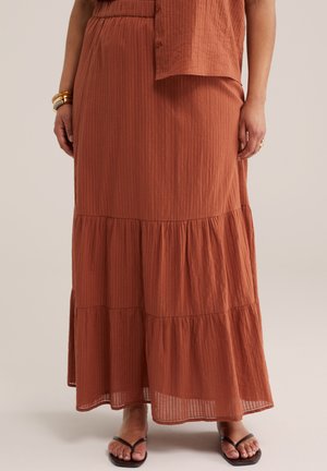 Femme portant une jupe longue à volants couleur rouille avec des rayures subtiles, associée à des sandales noires à bout ouvert et des bracelets en or.