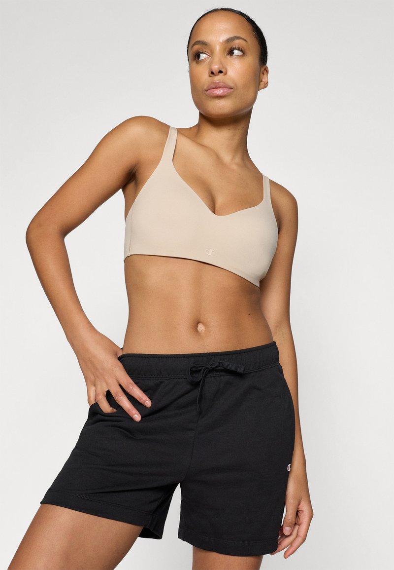 Top sportivo beige con scollo a V e spalline sottili, abbinato a pantaloni corti neri con coulisse e un piccolo logo sull'orlo.