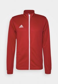 Veste de sport rouge en tissu lisse, dotée d'un col haut, d'une fermeture éclair blanche et d'un logo Adidas blanc sur la poitrine gauche.