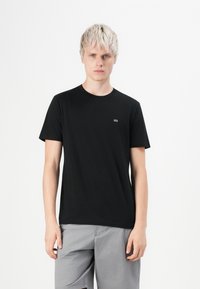 Junger Mann mit kurzen blonden Haaren, der ein schwarzes Kurzarm-T-Shirt und hellgraue Shorts vor einem schlichten weißen Hintergrund trägt.
