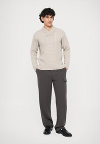 Μπέζ πουλόβερ πλεχτό με καλωδιοειδή σχέδια, γιακά σαλονιού, συνδυασμένο με σκούρες γκρι παντελόνες σε φαρδιά γραμμή και μαύρα παπούτσια.