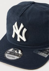 Gorra de béisbol azul marino con un logo bordado en blanco, visera curvada y correa ajustable; cuenta con una tela texturizada y una etiqueta circular de la marca en el ala.