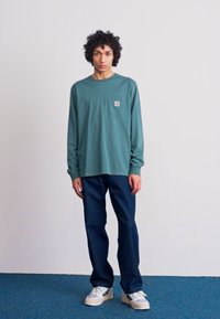 Camisa de algodón de manga larga en color verde azulado con un bolsillo en el pecho, combinada con jeans azul oscuro y zapatillas blancas. Fondo liso y suelo azul.