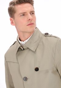 Gabardina beige de corte holgado, con cierre cruzado, grandes botones oscuros y cuello con solapas. Tejido liso y texturizado.