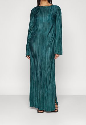 Kobieta ubrana w długą, plisowaną sukienkę w kolorze morskim z długim rękawem i wysokim kołnierzem oraz czarne sandały na paski.