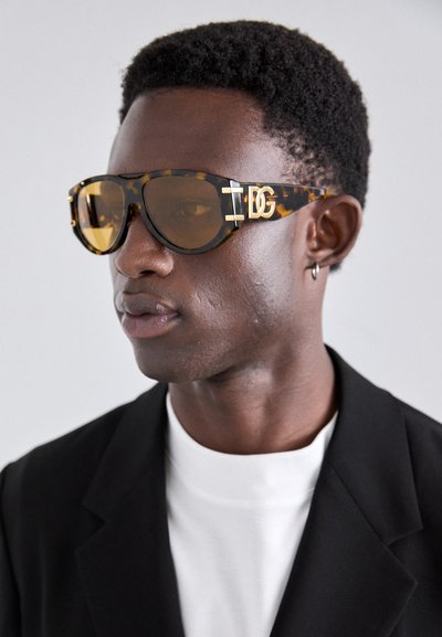 Ochelari de soare cu model țestoasă, cu lentile ambrate, formă rectangulară îndrăzneață, logo gravat pe tâmplă, asortați cu un sacou negru și o cămașă albă.