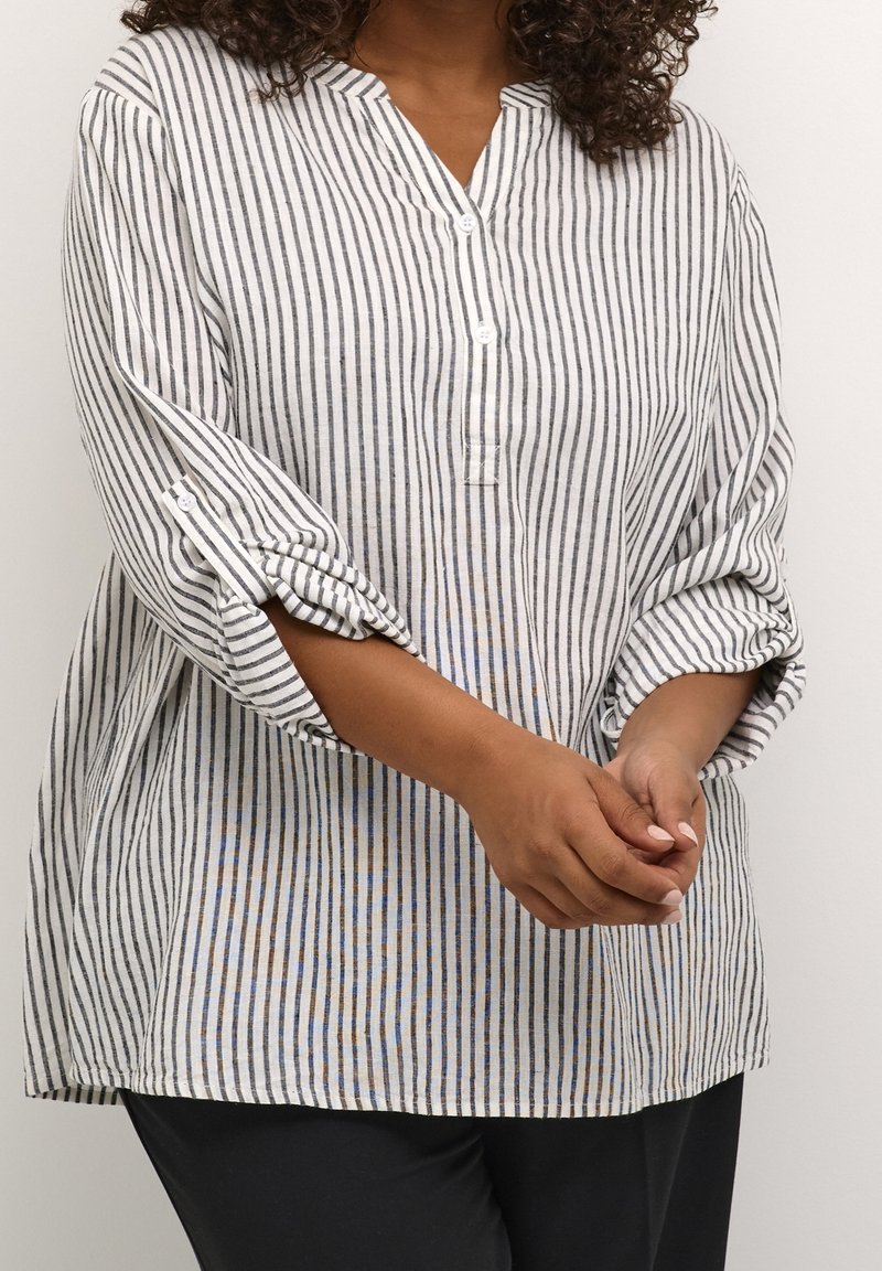 Personne portant une chemise blanche boutonnée à rayures noires verticales et manches retroussées, debout, mains jointes devant.