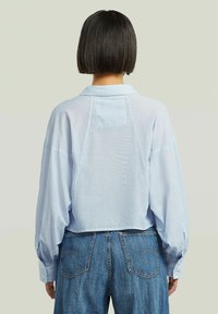 Camicia cropped azzurra chiara con colletto a punta, spalle scese e polsini abbottonati; tessuto morbido e strutturato con una vestibilità rilassata.