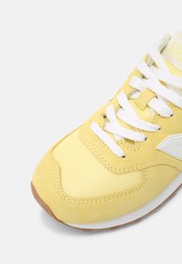 Zapatilla amarilla con materiales de ante y malla, que cuenta con cordones blancos, una superficie texturizada y una suela de goma. Los detalles incluyen un logo en el lateral.