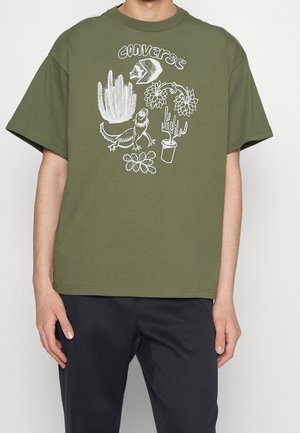 Camiseta de algodón verde oliva con ilustraciones gráficas blancas de plantas y una iguana, con un corte relajado y mangas cortas.
