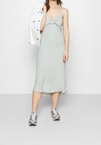 Robe midi à pois vert clair avec un détail à l'avant en torsion, des bretelles spaghetti ajustables et un ourlet volanté. Associée �à des baskets grises.