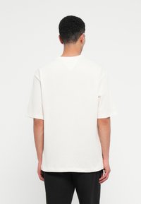 Άνδρας με κοντά σγουρά μαλλιά, ντυμένος με λευκό πουκάμισο με υφή και μαύρο παντελόνι, στέκεται με την πλάτη στραμμένη προς τον φακό σε λευκό φόντο.