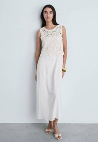 Langes weißes Kleid mit einem gehäkelten Spitzen-Oberteil, floralen Mustern und einem glatten Rock. Getragen mit hellen Sandalen und einem goldenen Armband.
