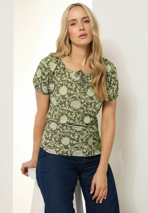 Green floral print top with puff sleeves, round neckline, and tie detail at the front. Worn with dark blue jeans.
