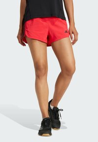 Shorts de sport rouges en tissu léger, avec un ourlet incurvé et un logo Adidas noir. Associés à des chaussures de course noires.