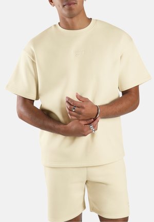 Man draagt een beige Reebok shirt met korte mouwen en bijpassende korte broek, houdt zijn handen samen waarbij ringen en een armband zichtbaar zijn.
