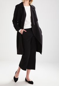 Eine Frau steht selbstbewusst in einem schwarzen Mantel über einem weißen Pullover und schwarzen weit geschnittenen Hosen, trägt schwarze spitze Schuhe vor einem hellen Hintergrund.