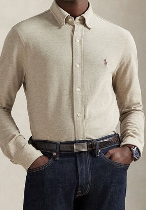 Mies, jolla on beige pitkähihainen napillinen paita tummansinisten farkkujen sisällä, musta vyö ja rannekello, kädet taskuissa.