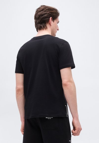 Άνδρας με καστανά μαλλιά που φοράει απλό μαύρο κοντομάνικο μπλουζάκι και μαύρα σορτς, γυρισμένος προς τα πίσω σε απλό φωτεινό φόντο.