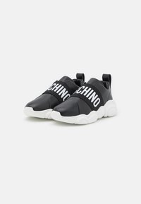 Baskets noires à enfiler en matériau synthétique lisse avec un grand logo blanc sur le dessus. Semelle en caoutchouc blanche épaisse.