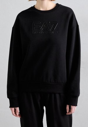 Osoba nosząca czarną, długorękawową bluzę z wytłoczonym logo "EA7 Emporio Armani" na piersi, w zestawieniu z czarnymi spodniami.