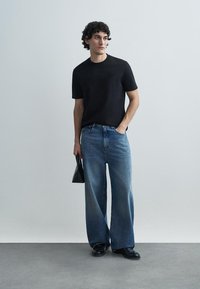 Svart T-shirt med korta ärmar, blå vida jeans, svarta skor, håller en svart väska, står på ett grått golv mot en ljus bakgrund.