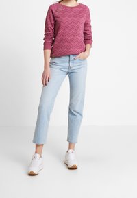 Persona vistiendo una camiseta de manga larga con un patrón de zigzag púrpura, jeans de color azul claro y zapatillas blancas, de pie contra un fondo blanco liso.