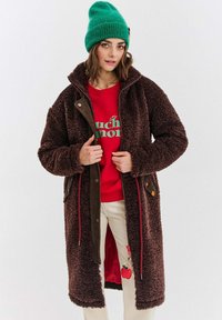 Žena v zelené pletené čepici, červené mikině, krémových kalhotách s klíčenkou jablka a dlouhém hnědém fleecovém kabátu, který drží otevřený.