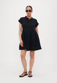 Rochie scurtă neagră cu mâneci scurte, cu un sistem de butoane pe față, în formă de A și talie strânsă. Pur tată cu sandale plate negre și ochelari de soare. Textura materialului este netedă.