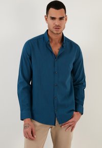 Camisa de botón de manga larga en color azul verdoso con cuello estructurado, botones oscuros y una textura suave, llevada sobre pantalones beige.