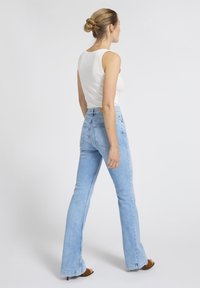 Vrouw met haar in een knot, gekleed in een witte mouwloze top, lichtblauwe high-waisted wijde spijkerbroek en bruine sandalen met hakken, zijwaarts staand.