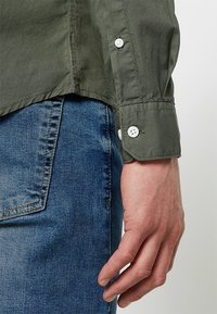 Oliwkowa koszula z długimi rękawami, z białymi guzikami i gładką fakturą. W połączeniu z niebieskimi dżinsami, prezentującymi swobodny styl.