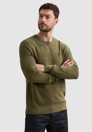 Man with short brown hair and beard wearing an olive green knit sweater with textured stripes, arms crossed, looking slightly to the side.