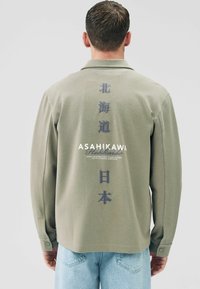 Olive green long-sleeve polo shirt with textured fabric, featuring vertical Japanese text on the back and brand name in white.