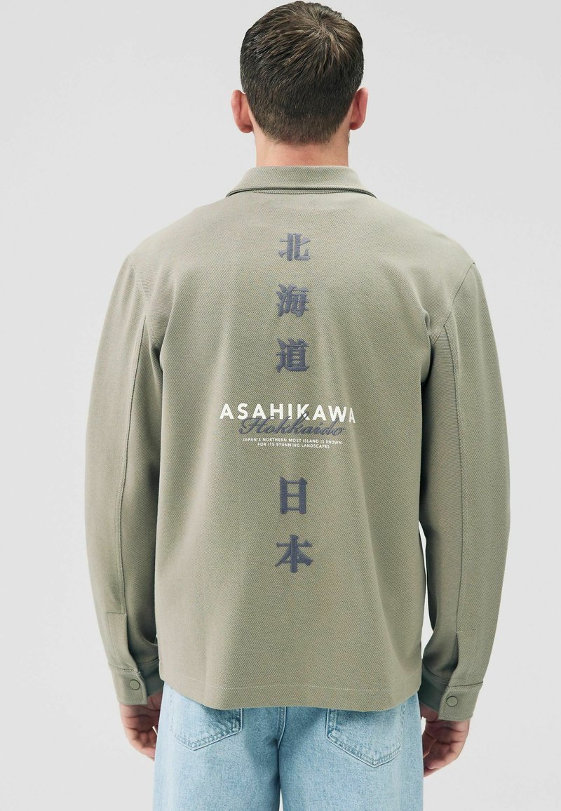 Olive green long-sleeve polo shirt with textured fabric, featuring vertical Japanese text on the back and brand name in white.