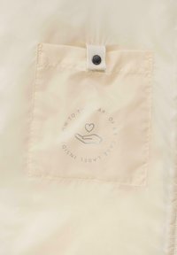 Hellbeige Stofftasche mit einem Druckknopfverschluss. Pflegeetikett im Inneren mit einer Hand- und Herzgrafik, umgeben von Pflegehinweisen.