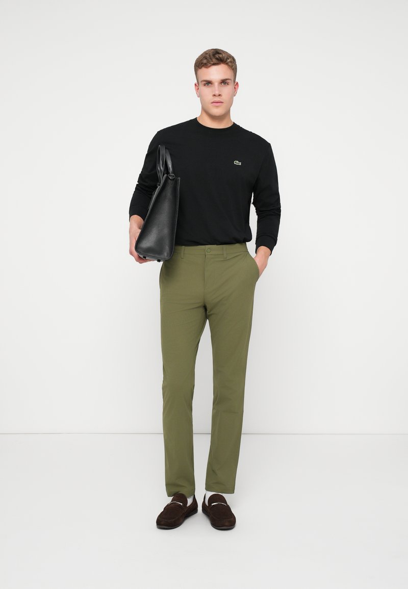 Camicia nera a maniche lunghe con logo verde, pantaloni verde oliva e scarpe scure senza lacci. Il modello tiene in mano una borsa nera.