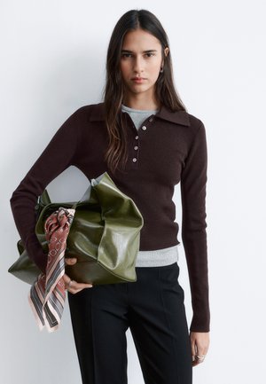 Kobieta o długich ciemnych włosach, ubrana w ciemnobrązowy sweter i czarne spodnie, trzymająca dużą zieloną torebkę z wzorzystą chustą.