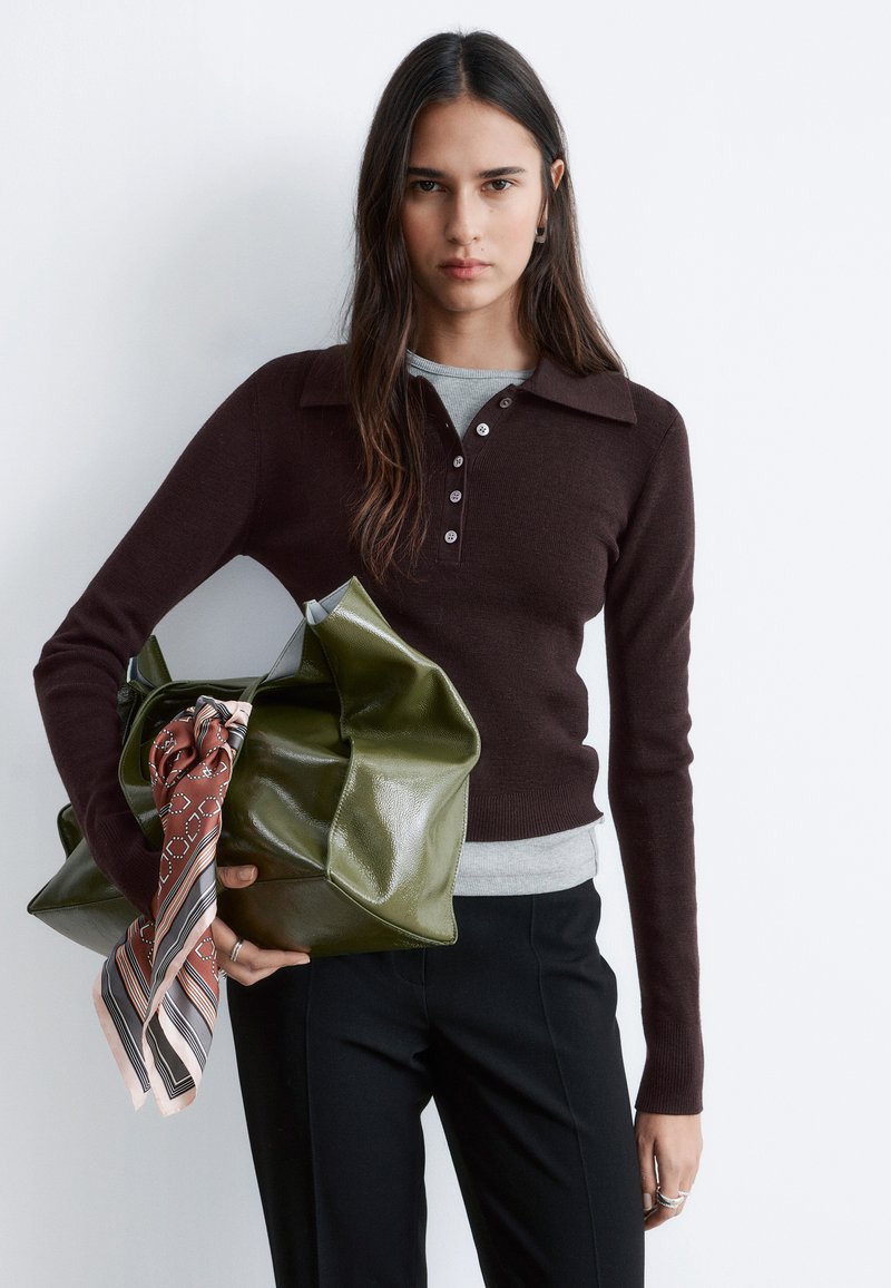 Frau mit langen dunklen Haaren, die einen dunkelbraunen Pullover und schwarze Hosen trägt, hält eine große grüne Handtasche mit einem gemusterten Schal.