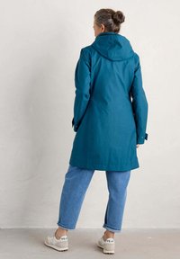 Tealfärgad regnjacka med huva, med en rak passform, knappar på ärmarna, tillsammans med ljusblå jeans och vita sneakers.