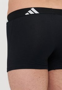 Črne boksarske hlače iz mehkega, raztegljivega materiala. Imajo širok elastičen pas z belim logotipom Adidas in diagonálnimi črtami.