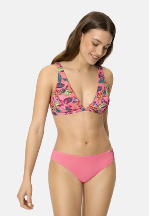 Mujer sonriente con un bikini rosa, parte superior con estampado floral y parte inferior lisa rosa, posando contra un fondo claro y uniforme.